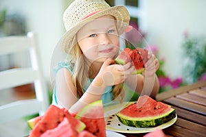 Funny little girl wearing straw hat biting a slice of watermelon outdoors on warm and sunny summer day.