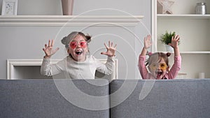 Funny little girls play at home, jump out from sofa, scare, scream and laugh, look at camera