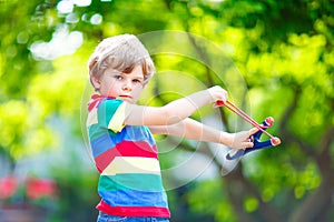 Funny little kid boy shooting wooden slingshot against green tree background. Child having fun in summer