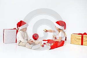 Funny little kids in Santa hat sitting between gift boxes and playing with christmas balls. Isolated on white background. New year