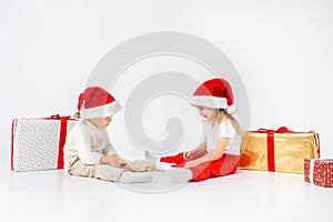 Funny little kids in Santa hat sitting between gift boxes and playing with christmas balls. Isolated on white background. New year