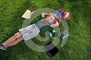 Funny little school child with digital gadgets and backpacks sitting at the grass park outdoors. Top view of little boy