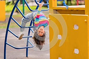 Funny little smiling girl 5-6 years old, hanging upside down on the children& x27;s stairs at the playground