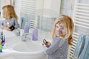 Funny little toddler girl using mother& x27;s make up and painting face with eye shadows. Happy baby child making