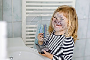 Funny little toddler girl using mother`s make up and painting face with eye shadows. Happy baby child making experiments