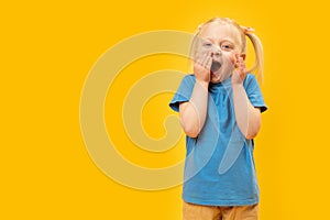 Funny little white-haired girl with two ponytails in blue T-shirt. Yellow background. Child laughs by covering his mouth with her