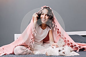 Funny lovely home moments of happy young woman in pajama under pink blanket playing with little white dog on bed in