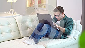 Funny male in glasses is typing on laptop on sofa