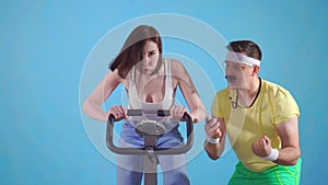 Funny man coach of 80`s with mustache trains a young woman on exercise bike on a blue background