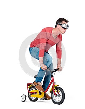 Funny man in goggles, jeans and a red t-shirt pedals a children`s bicycle, isolated on white background.