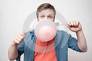 Funny man holding in his mouth a balloon and going to burst it with a knife and fork. White background.