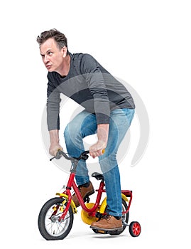 Funny man in jeans and black t-shirt pedals a children`s bicycle, isolated on white background.