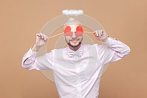 Funny man with nimb over head, standing covering his eyes with little red hearts, hiding.
