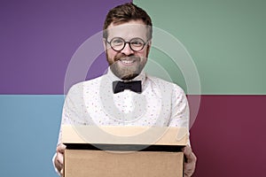 Funny man in round glasses holds a box with a gift in hands, he smiles and looks joyfully at the camera.