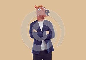 Funny man in rubber horse mask standing with crossed on chest hands on beige background.