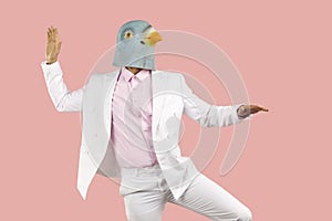 Funny man in rubber pigeon mask and in white formal suit dancing on pink background.