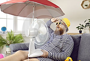 Funny man suffering from summer heat, sitting at home, sweating and holding electric fan