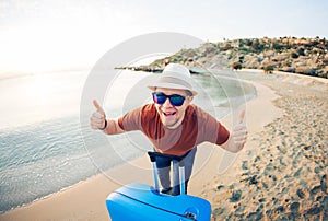 Funny man with suitcase showing thumbs up against the blue ocean. Travel concept.