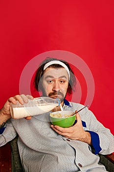 Funny mature bearded man wearing homewear, pijamas has breakfast over bright red background. Positive emotions, home