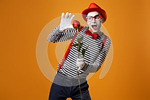 Funny mime with white face in red hat and striped t-shirt holding rose isolated on empty orange background