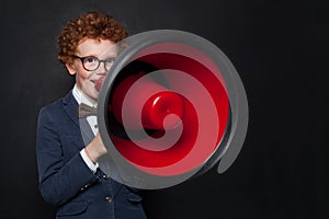 Funny mischievous child boy speaking through a loudspeaker on blackboard background. Kid with red megaphone