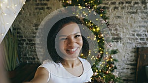 Funny mixed race girl taking selfie pictures on smartphone camera at home near Christmas tree