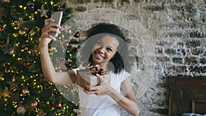 Funny mixed race girl taking selfie pictures on smartphone camera at home near Christmas tree