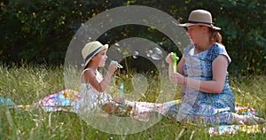 Funny mom and little daughter blow soap bubbles sitting on the lawn in the park