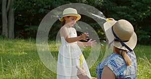 Funny mom and little daughter blow soap bubbles sitting on the lawn in the park