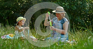 Funny mom and little daughter blow soap bubbles sitting on the lawn in the park