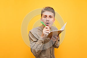 Funny nerd in glasses and shirt looks surprised at the camera and writes in a notebook on a yellow background. Portrait of funny