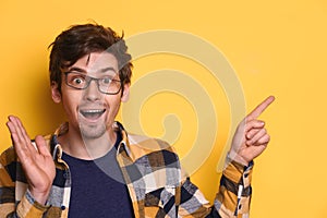 Funny nerd guy looks amazed and shows something on yellow studio wall