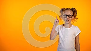Funny nerd schoolgirl raising finger up isolated on orange background, good idea