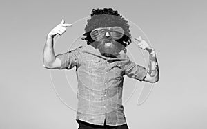 Funny nerdy guy in funny goggles pointing finger. Crazy funny bearded man with wig and fun glasses on sky background