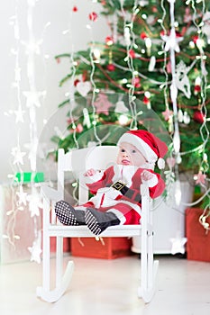 Funny newborn baby boy in Santa costume sitting under Christmas tree