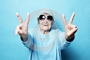 Funny old lady wearing blue sweater, hat and sunglasses showing victory sign. Isolated on blue background.