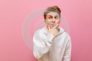 Funny pensive guy in white casual clothes stands on a pink background, looks away at a blank spot with a funny face. Ceerful young