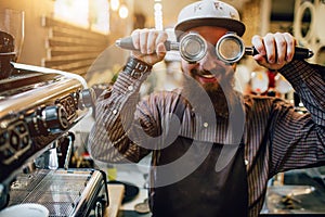 Funny picture of bearded barista in apron holding tow cezve and cover eyes with them. He smiles a bit. Guy stand at