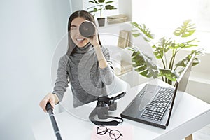 Funny picture of a girl sitting at the table in a big bright room and recording video. She is looking to video camera