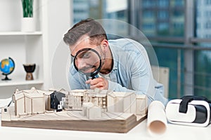 Funny picture of male architect looks through magnifying glass to a house model. House inspection and real estate