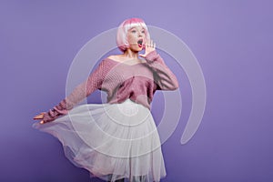 Funny pink-haired girl in periwig posing in studio. Adorable female model wears white skirt and bright peruke enjoying