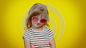Funny playful blonde kid child wearing sunglasses, looking with charming smile on yellow background