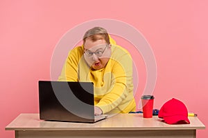 Funny plump man geek in yellow sweatshirt using laptop with funny freak grimace