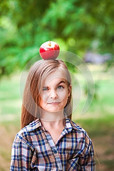 Funny portrait of a beautiful baby girl with a red apple on her head. A girl in a plaid shirt on a farm harvesting apples smiles a