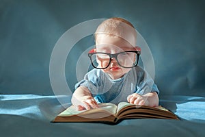 Funny portrait of cute baby in glasses. The baby lies on his stomach and reads an old book on a blue background