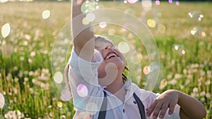 funny portrait of a five year old boy stands on a field of dandelions and catches soap bubbles from a toy gun at sunset