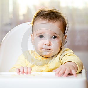 Funny portrait of a kid on a high chair indoors. The toddler looks at the camera. The child ate and smeared his face. Handsome