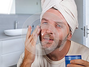 Funny portrait of young happy and attractive camp gay man in bathroom applying moisturizer facial cream with head wrapped in towel