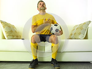 Funny portrait of young man in football team uniform watching soccer game on TV at home couch listening national anthem with hand