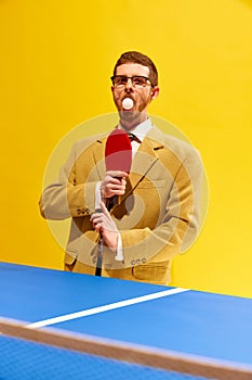 Funny portrait of young man in glasses and stylish suit standing with tennis ball inside mouth against bright yellow